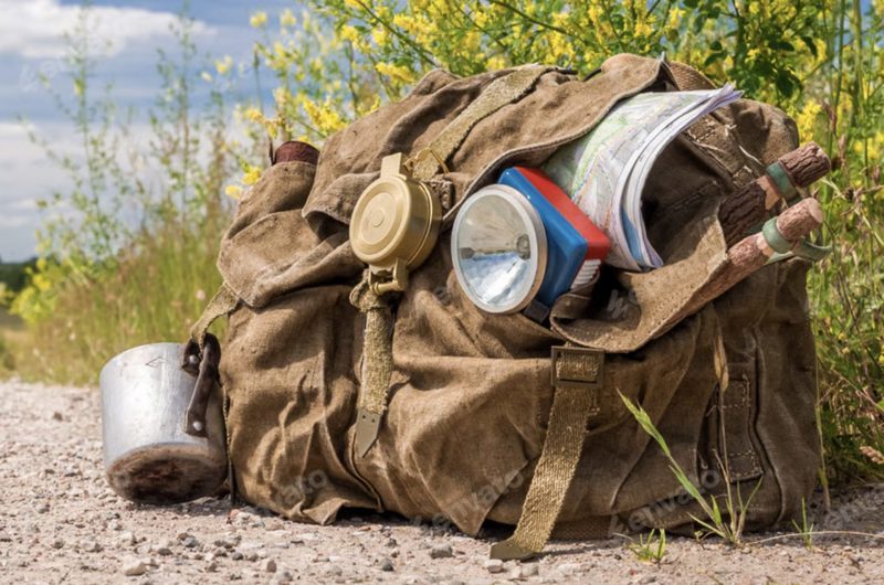 A heavily overloaded vintage canvas rucksack on a gravel roadside, hung with a compass, torch, map and cooking pot