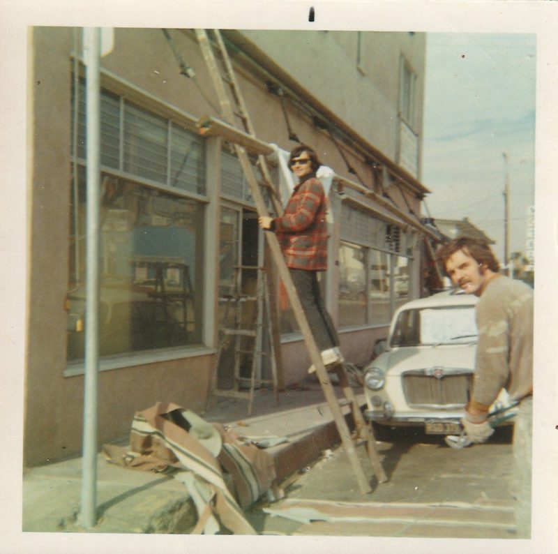 Two young men fitting a storefront window at Scott's 1970s Manhattan Beach laundromat, an old car parked alongside