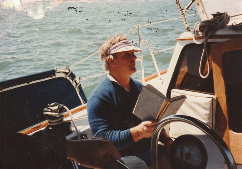Scott in a visor, reading a book at the wheel of his 38-foot sailboat