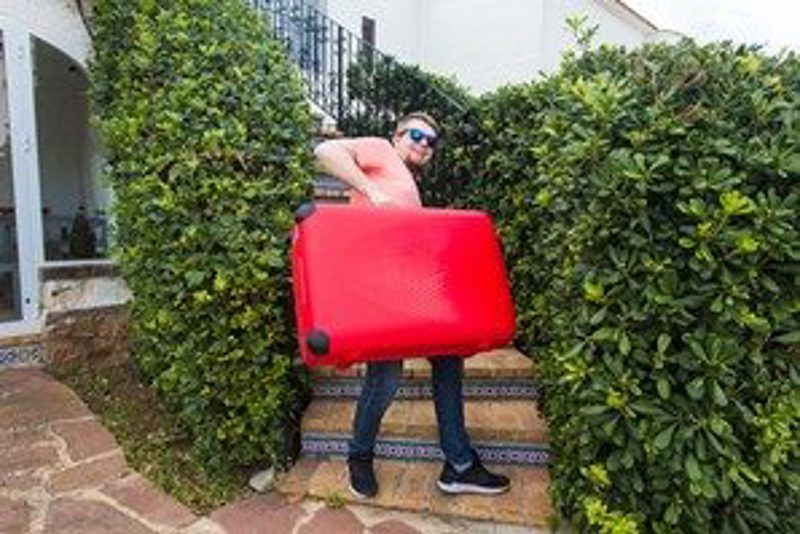 A man in sunglasses hoisting an oversized red hard-shell suitcase up a flight of steps