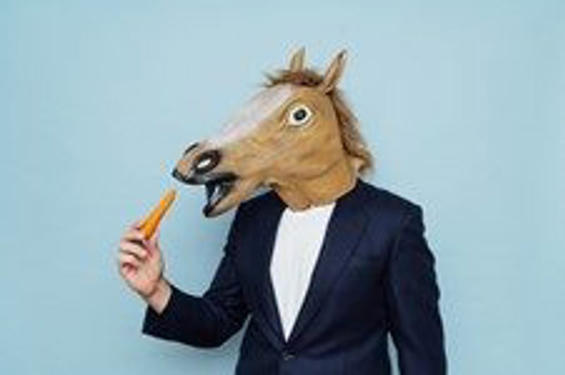 A man in a dark suit wearing a horse mask, holding a carrot