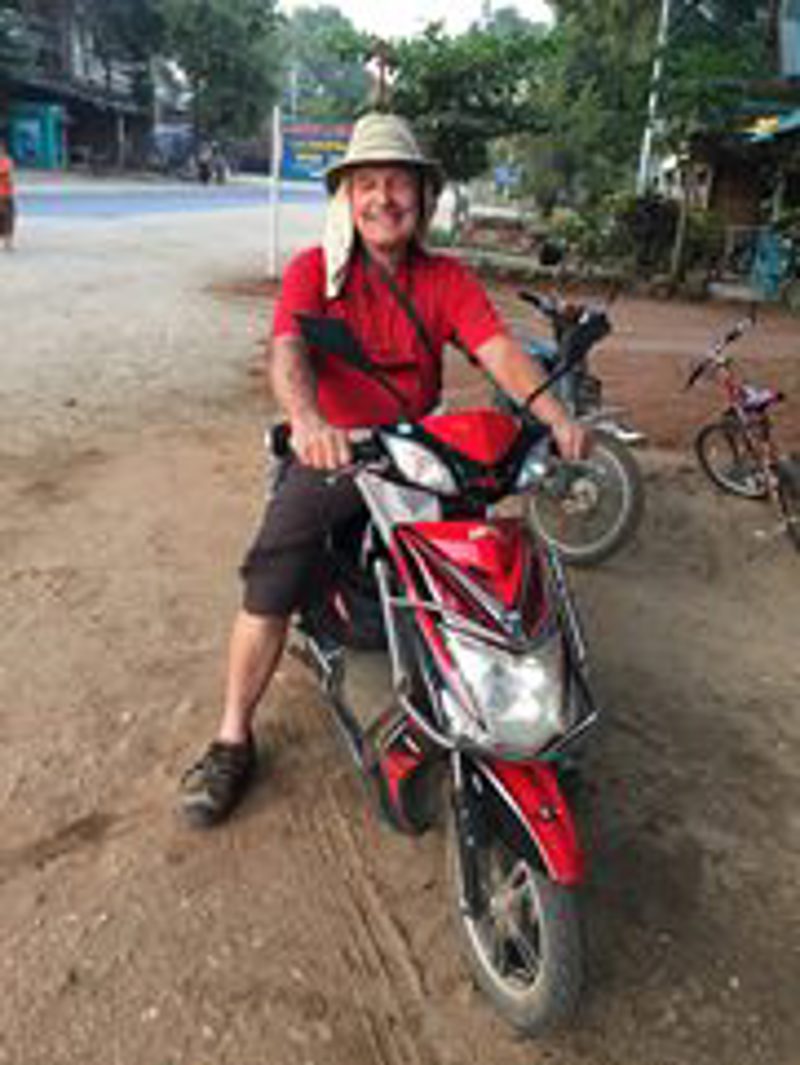 Scott in a wide-brimmed hat, seated on a rented red scooter on a dirt village road in Myanmar